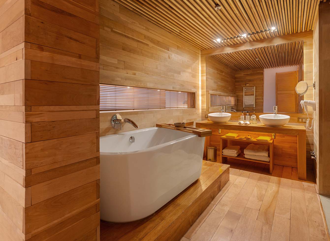 Spacious bathroom with wood paneling throughout, white soaking tub, dual vessel sinks on wooden counter, and recessed lighting in slatted ceiling.