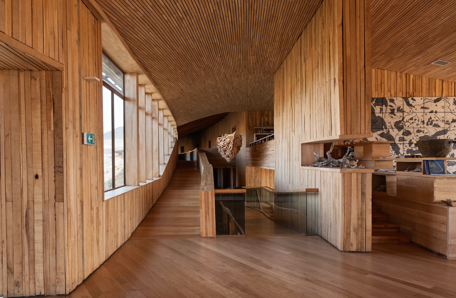 Interior corridor with wood paneling, large windows, display shelves, and a sculptural hanging element in a modern lodge setting.