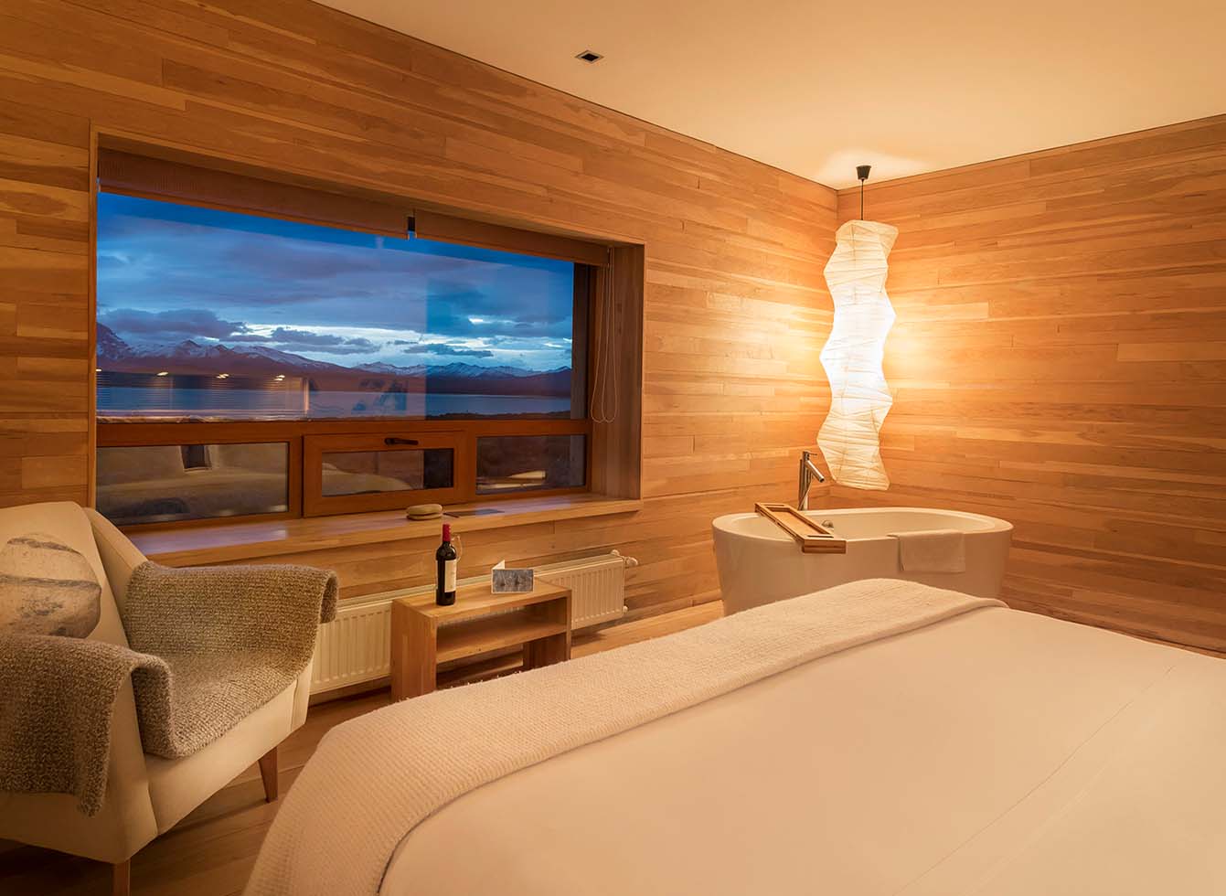 Modern lounge with warm wood ceiling, orange and olive seating areas, pendant lights, and a large cone-shaped fireplace with guests scattered throughout.