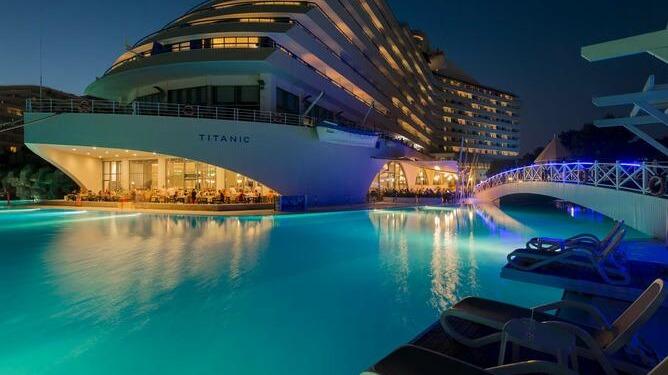 Titanic Deluxe Lara's illuminated pool complex at dusk, with turquoise water, loungers in foreground, and modern resort buildings lit in blue.