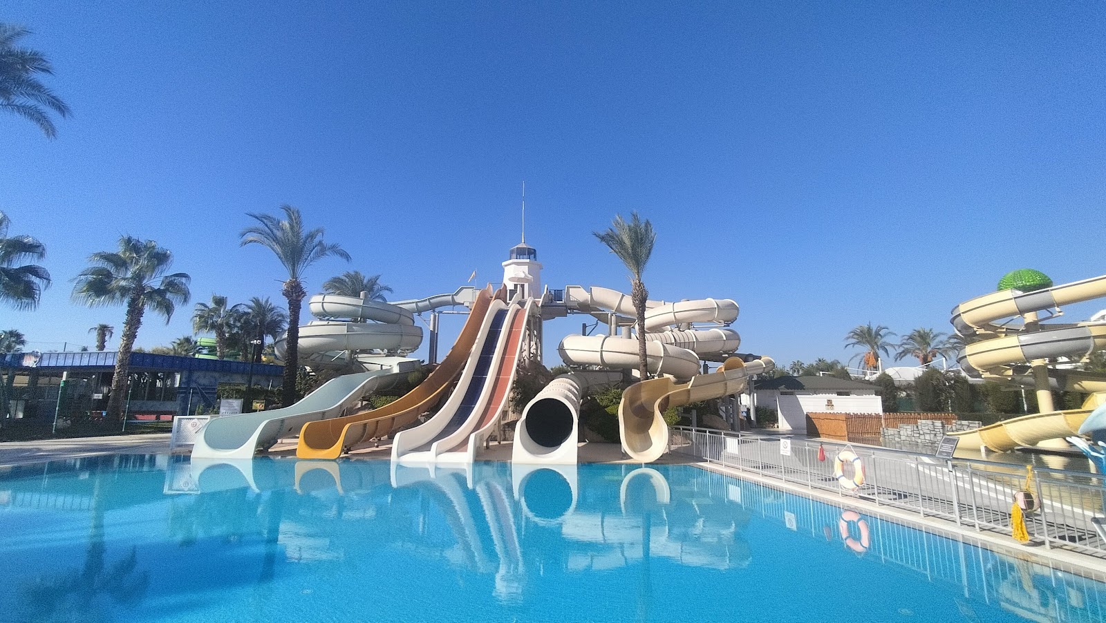 Water slides in multiple colors tower over a bright blue pool at Titanic Deluxe Lara, with palm trees and clear sky visible behind.