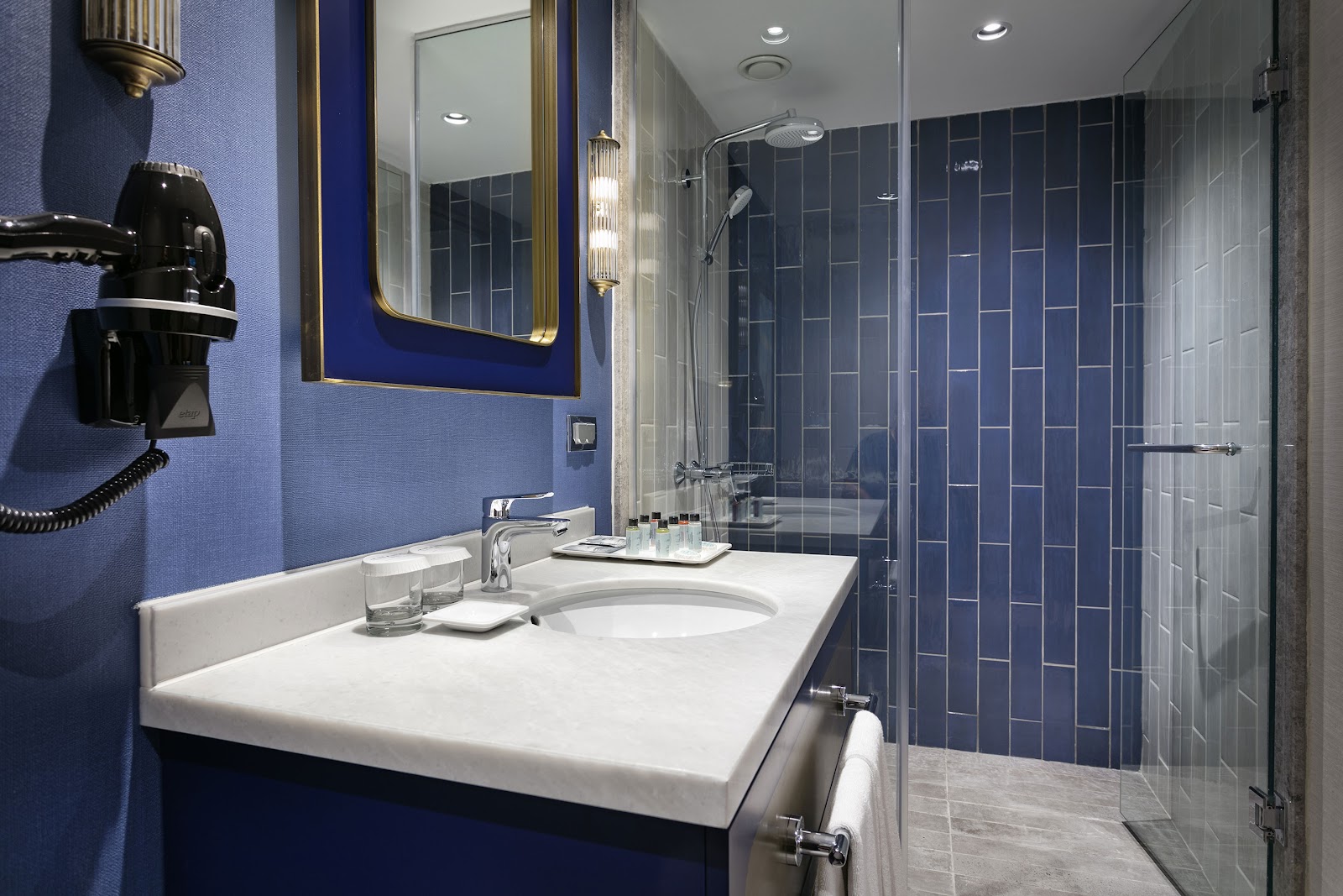 Modern bathroom with periwinkle walls, white vanity, brass-framed mirror, and walk-in shower with dark blue tile at Titanic Deluxe Lara.