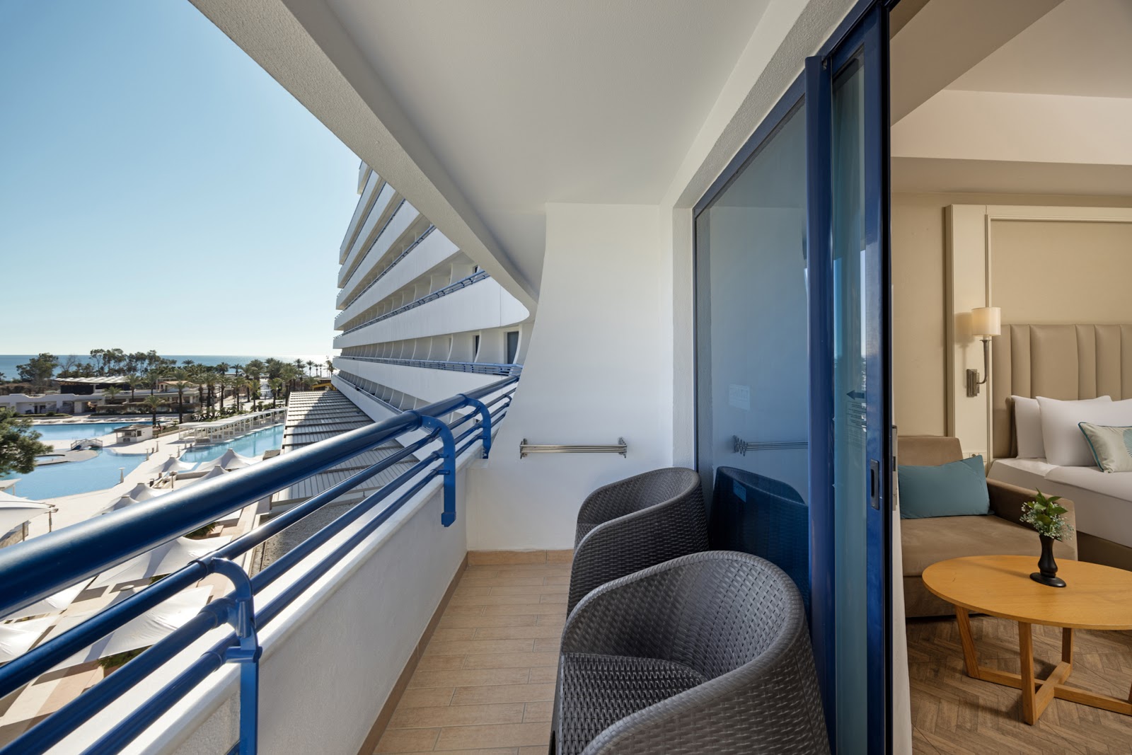 Resort room balcony with blue railing, two gray chairs, and view of pools, beach, and palm trees below.