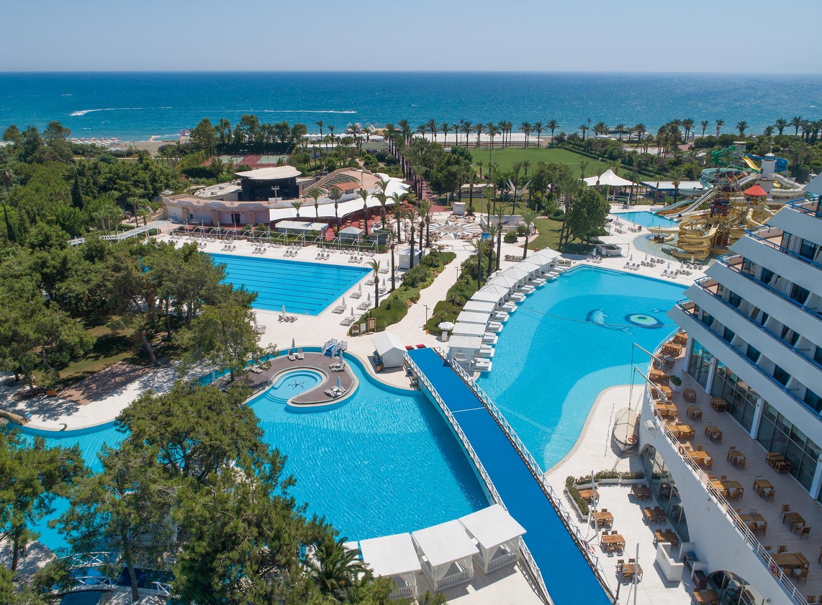 Aerial view of Titanic Deluxe Lara with multiple blue pools, sandy beach, palm trees, water slides, and Mediterranean Sea in the background.