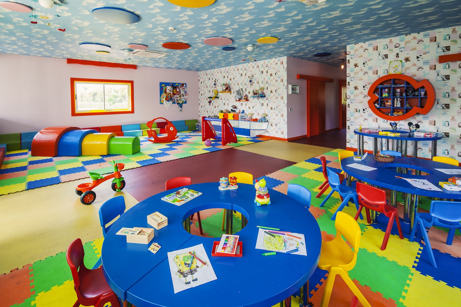 Colorful children's play room with blue round tables, foam tunnels, toy vehicles, and a sky-themed ceiling at Titanic Deluxe Lara.