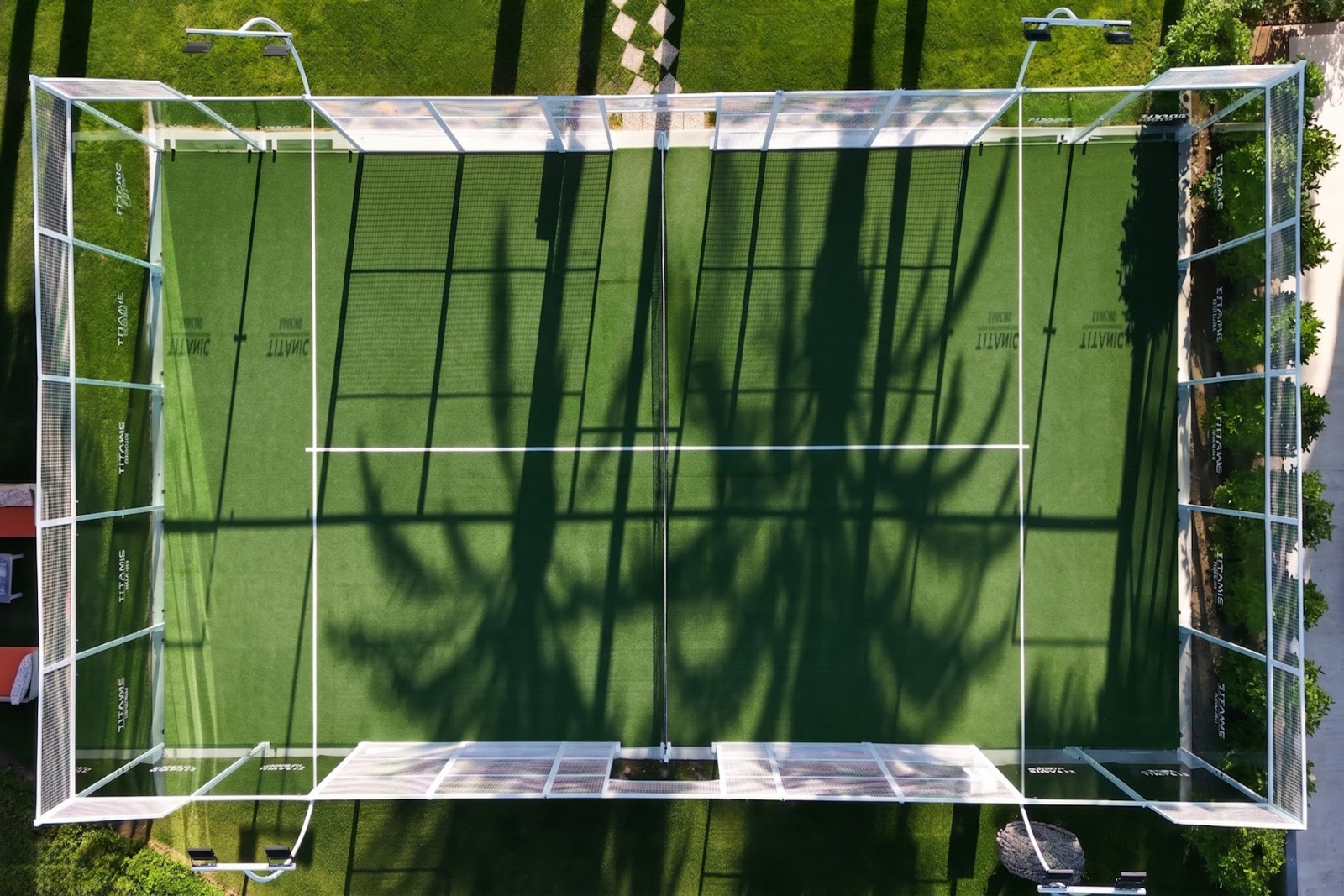 Aerial view of a padel tennis court on manicured grounds with palm trees, beach structures, and turquoise Mediterranean Sea in the background at Titanic Deluxe Lara.