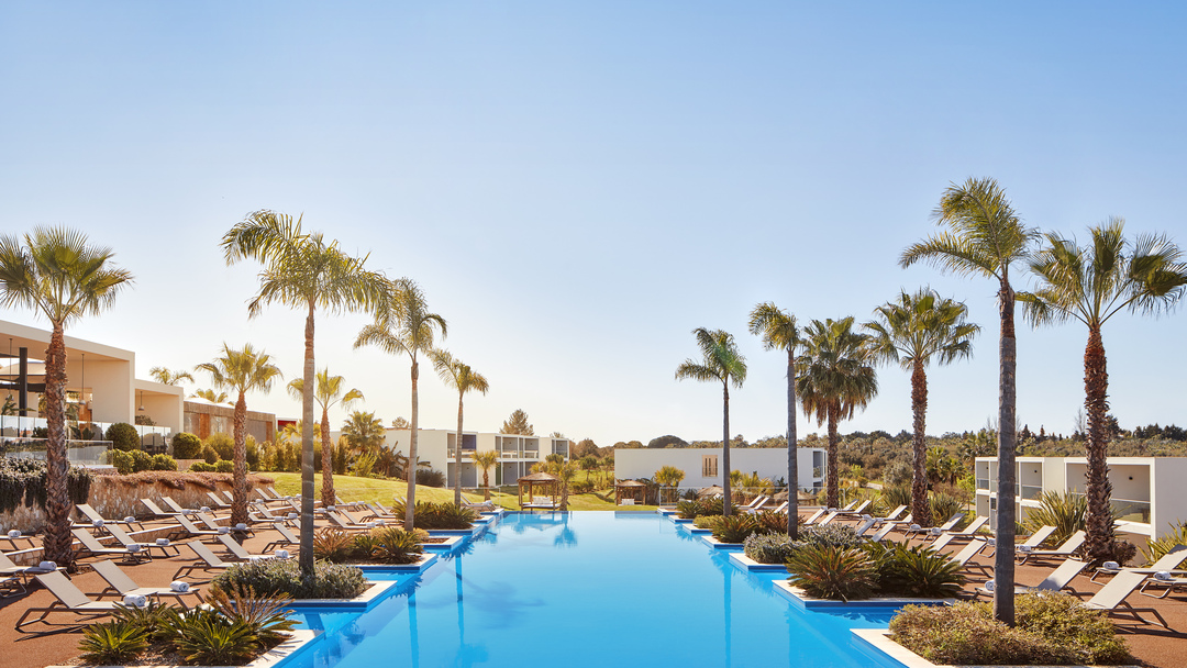 Long rectangular pool surrounded by loungers and palm trees at Tivoli Alvor Algarve, with resort buildings and clear sky beyond.