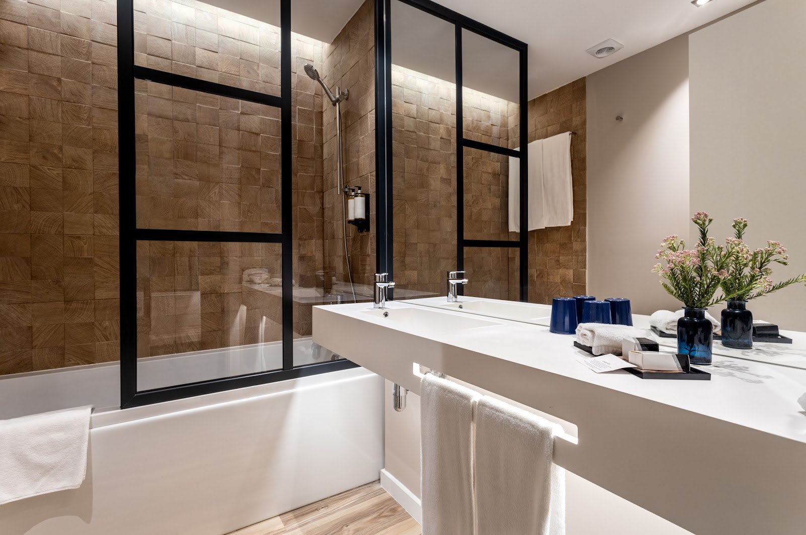 Modern bathroom with dual vanity sink, brown tile walls, black-framed glass shower enclosure, and white towels and amenities on counter.
