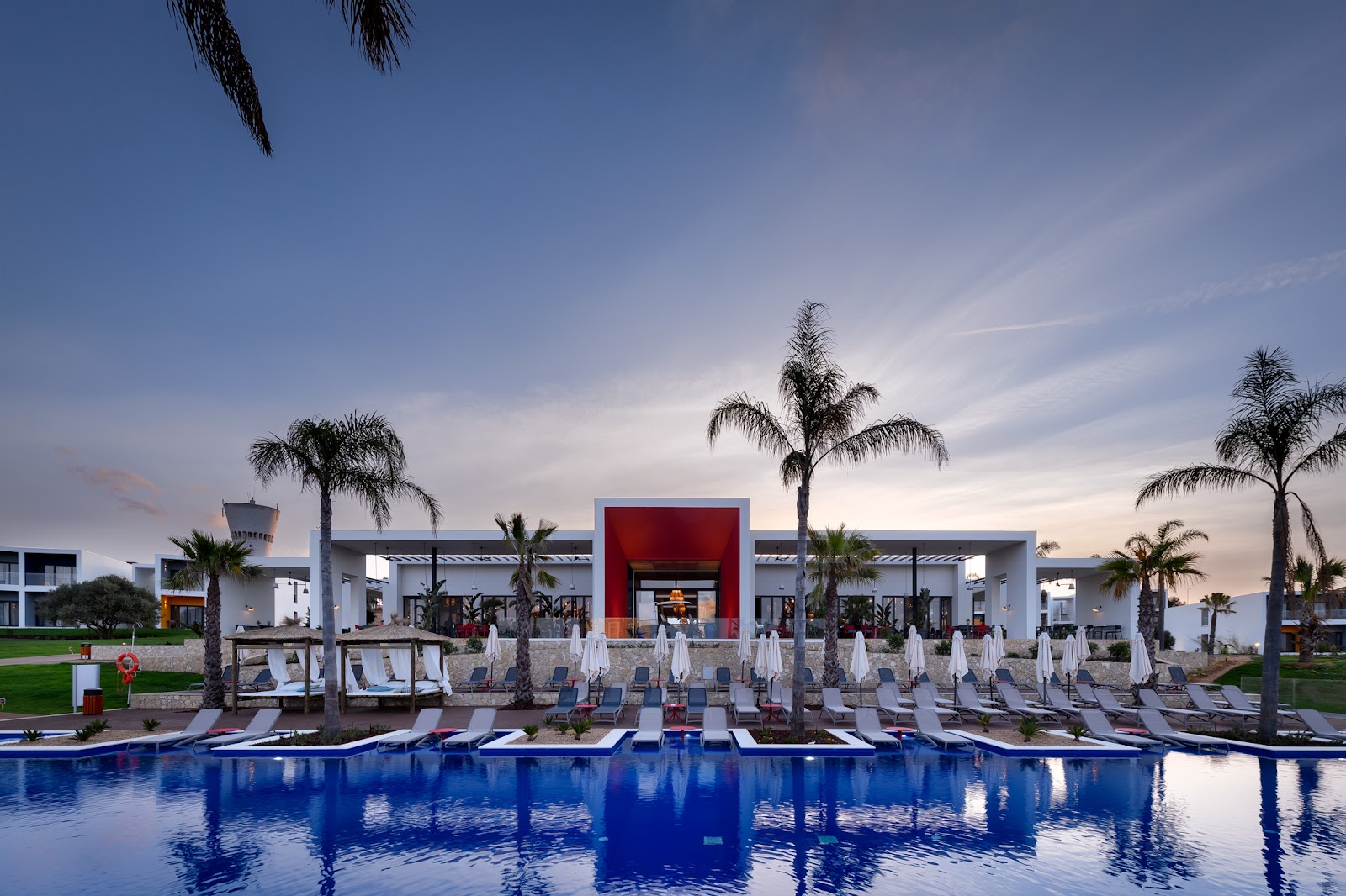 Large pool with white covered loungers facing a modern white resort building with red accent wall, palm trees, and manicured lawn at dusk.