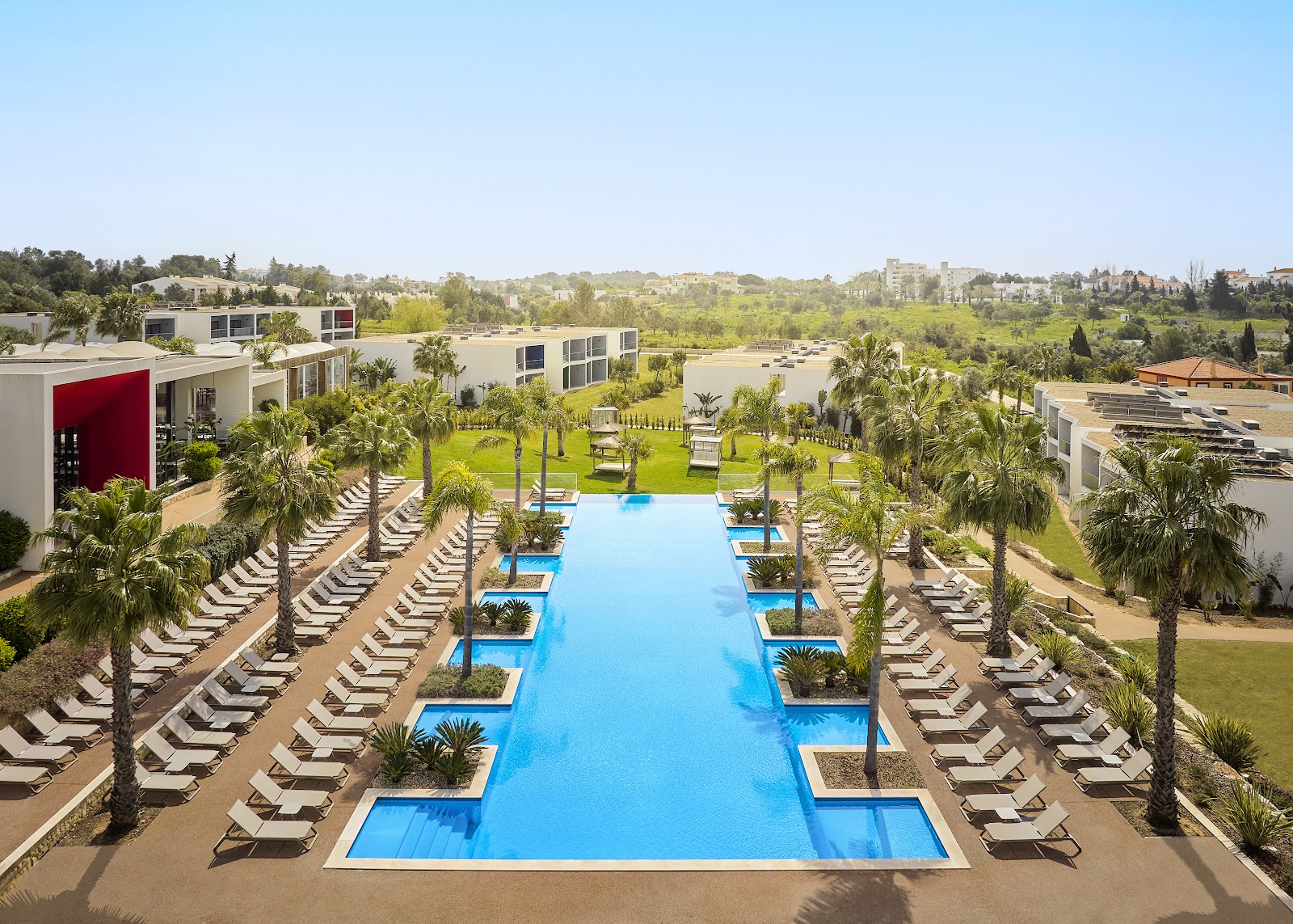 Long rectangular blue pool lined with rows of loungers and palm trees, modern white resort buildings and manicured lawns beyond.