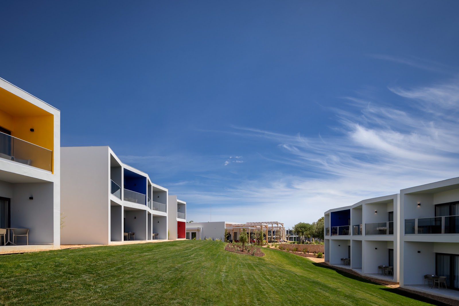 Modern white resort buildings with yellow and blue accents line a sloping green lawn under blue sky at Tivoli Alvor Algarve.