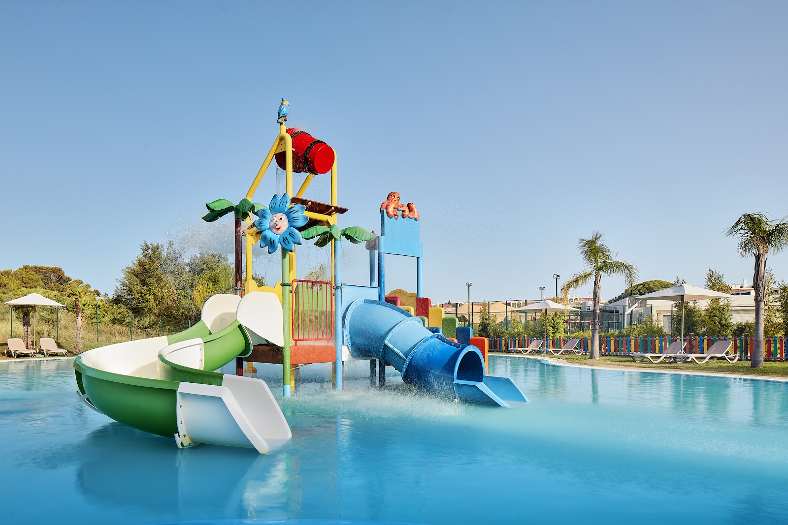 Colorful children's water play area with boat-shaped slide, blue tunnel slide, and climbing structures in shallow blue water at Tivoli Alvor Algarve.