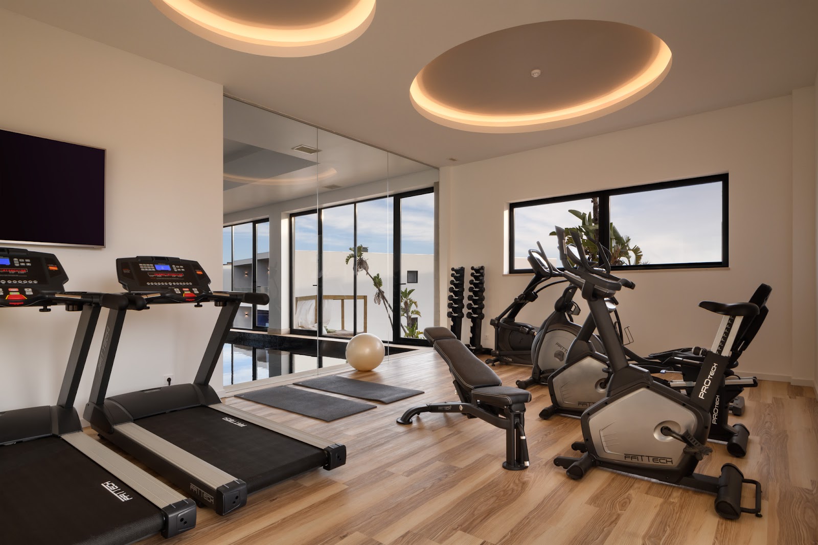 Modern fitness center with treadmills, stationary bikes, and exercise equipment under circular ceiling lights, overlooking the ocean through floor-to-ceiling windows.