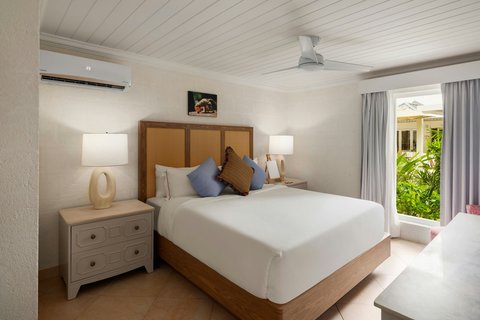 Bright bedroom at Treasure Beach Art Hotel with wooden bed, white linens, blue pillows, nightstand lamps, and open doors to tropical garden.