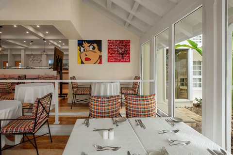 Bright dining area at Treasure Beach Art Hotel with white tablecloth table, plaid cushioned chairs, colorful wall art, and ocean view through glass doors.
