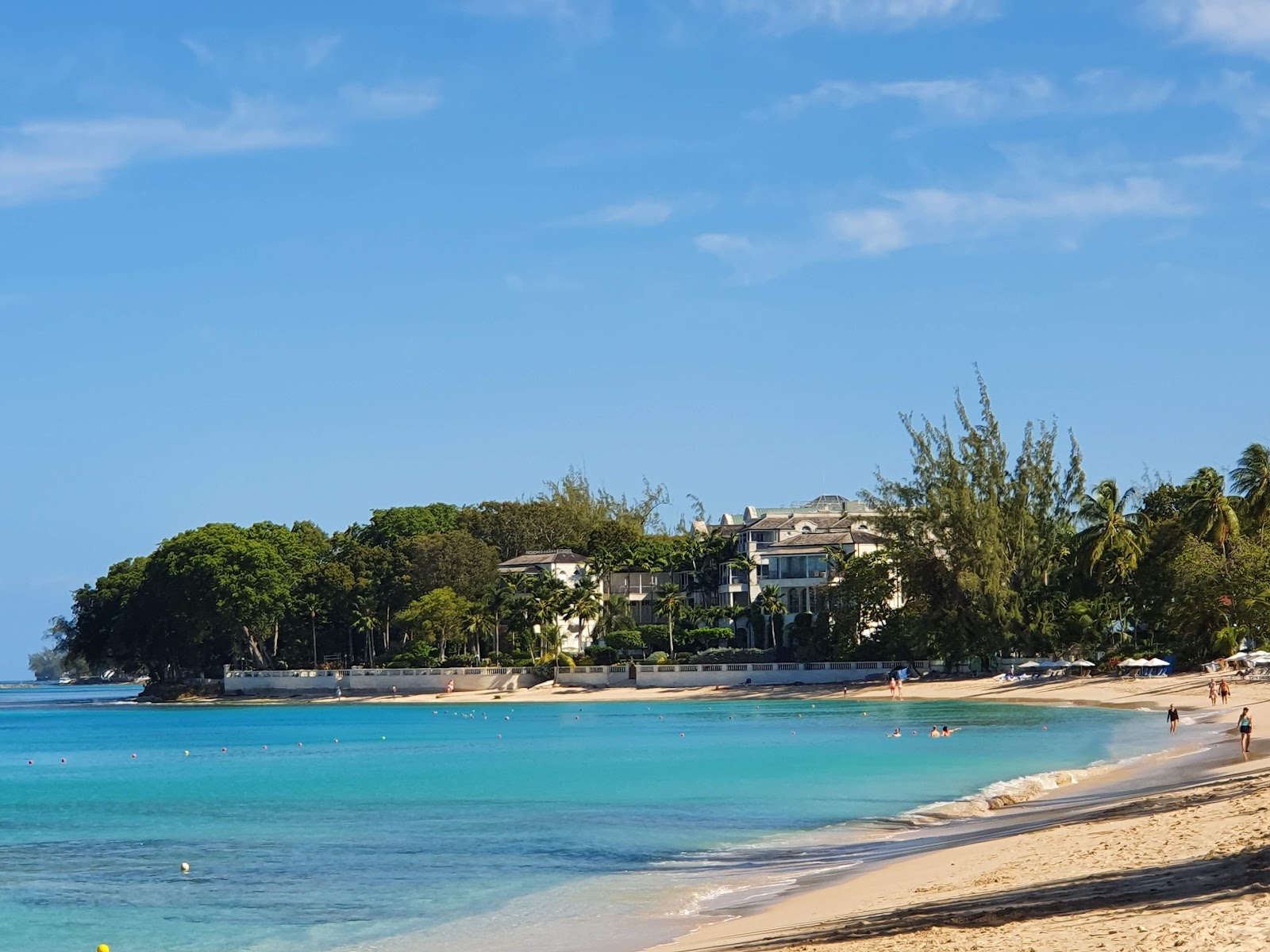 Treasure Beach Art Hotel sits on a sandy beach with turquoise water and swimmers, backed by lush tropical vegetation under clear blue sky.