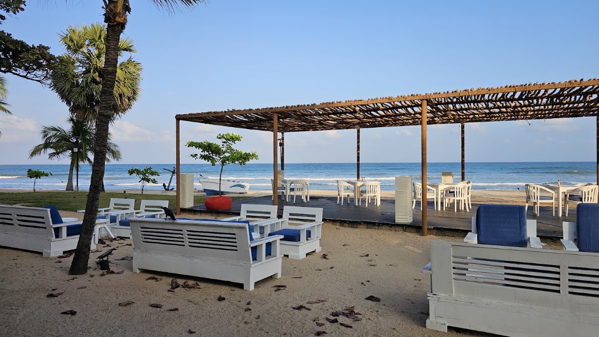 Beach cabanas with white and blue cushioned loungers sit on sand at Trinco Blu by Cinnamon, with a wooden pergola, palm trees, and ocean beyond.