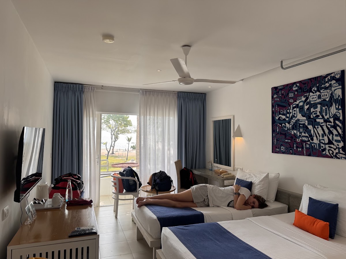 Guest room at Trinco Blu by Cinnamon with two beds, a guest resting on one bed, and a balcony overlooking the grounds and ocean beyond.
