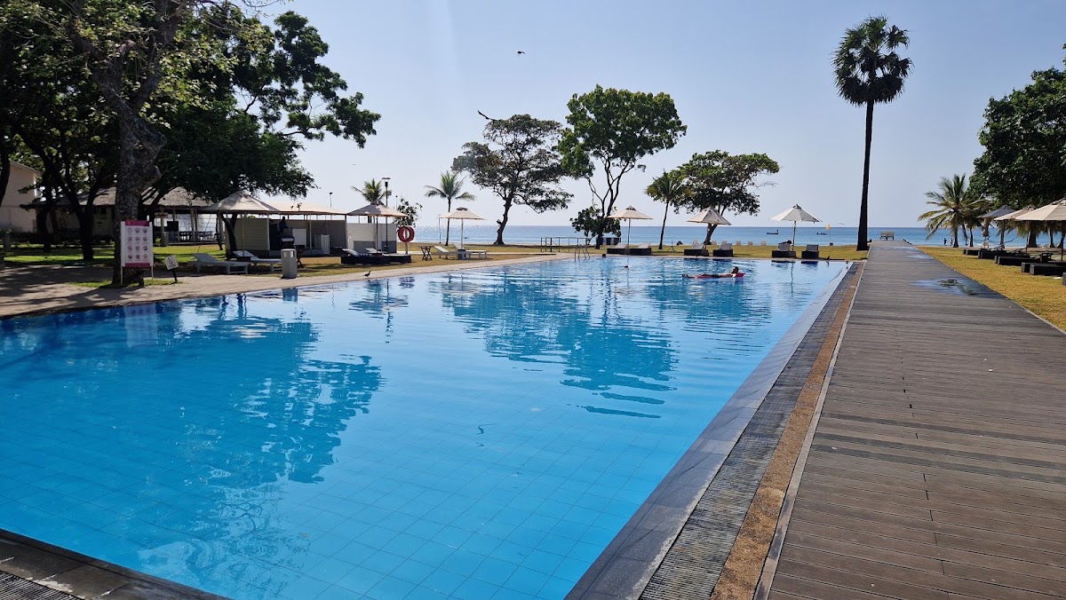Large resort pool with wooden deck overlooking the Indian Ocean, surrounded by palm trees and beach cabanas at Trinco Blu by Cinnamon.