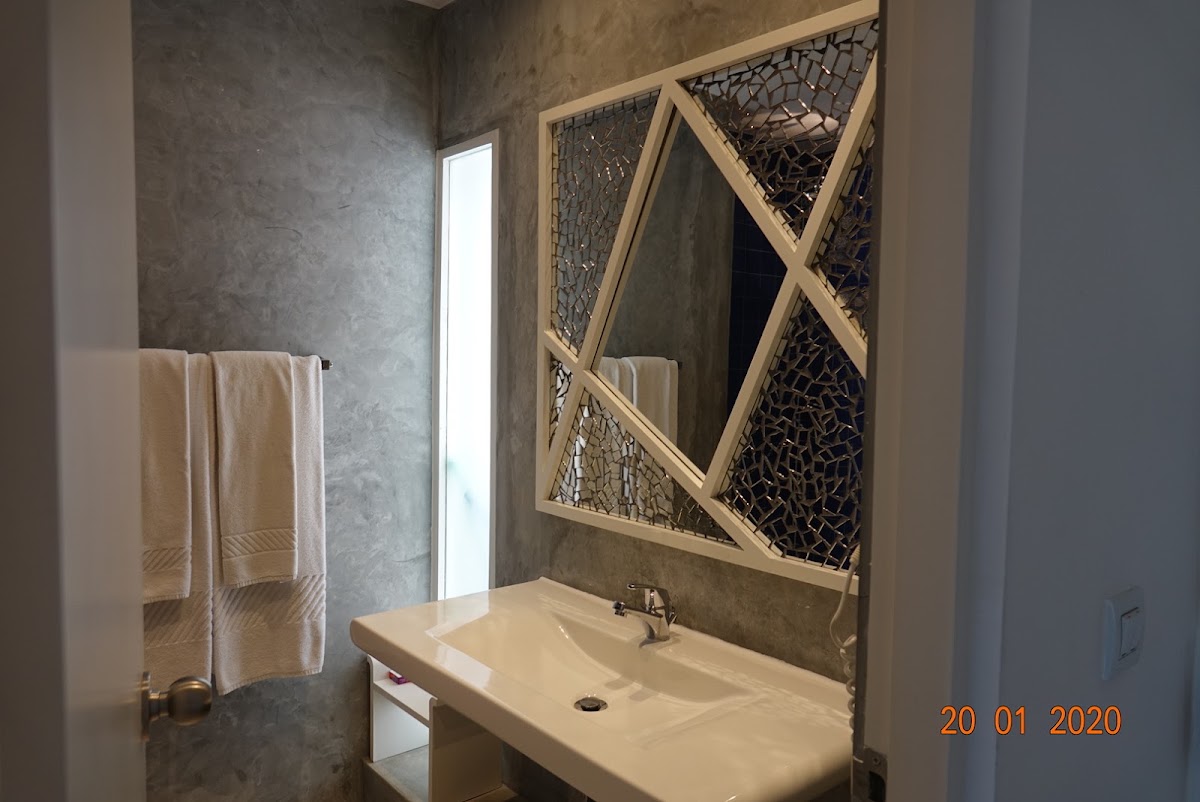Bathroom with concrete walls, white sink, wooden X-frame mirror with woven backing, and cream towels on rack.