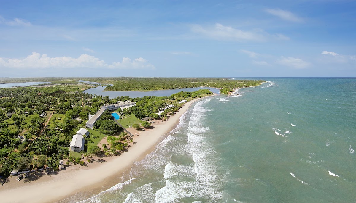 Aerial view of Trinco Blu by Cinnamon resort stretching along a sandy beach with turquoise waves, surrounded by tropical vegetation and lagoons.