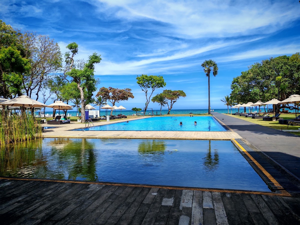 Trinco Blu by Cinnamon's beachfront pool with wooden deck, lounge chairs, umbrellas, and ocean view beyond palm trees under blue sky.