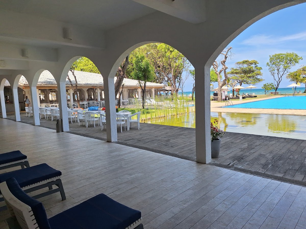 Covered terrace with white columns at Trinco Blu by Cinnamon opens to pool, lagoon, and ocean beyond with scattered trees.