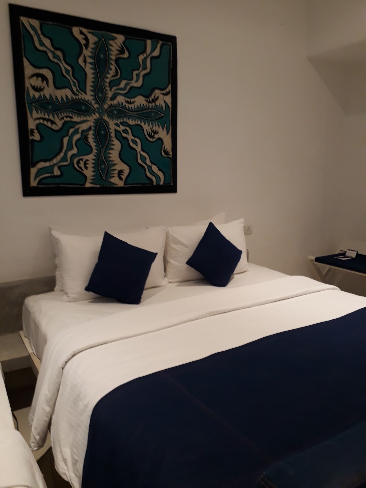 Resort bedroom with white and navy bedding, decorative navy pillows, and teal and cream patterned artwork on the wall above the bed.