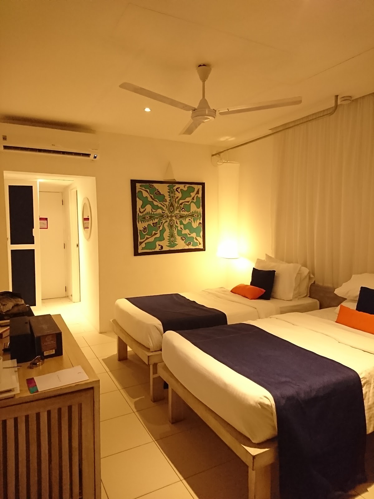 Hotel room with two white beds with dark runners, orange accent pillows, tropical artwork on wall, ceiling fan, and tiled flooring.