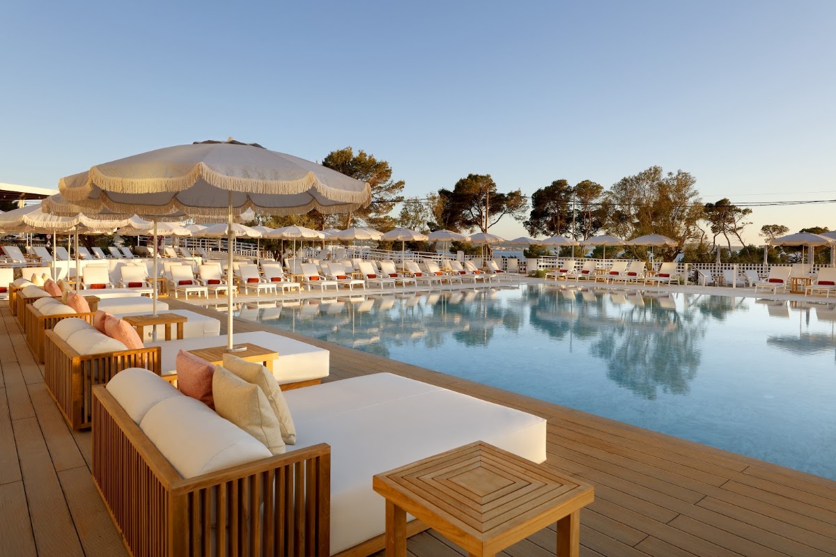 Wooden lounge chairs with cream cushions line a wooden deck overlooking a calm blue pool surrounded by cream umbrellas and sunbeds at TRS Ibiza.