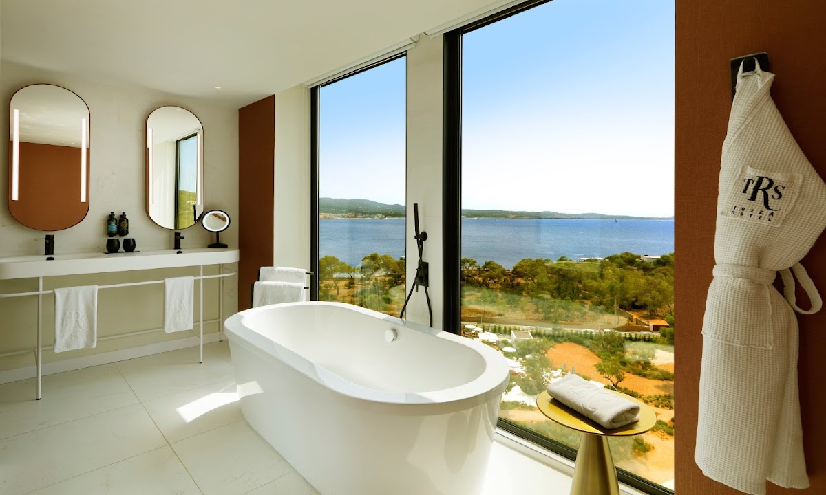 Modern bathroom with freestanding white tub, double vanity, and floor-to-ceiling windows overlooking Mediterranean coastline and forested landscape.