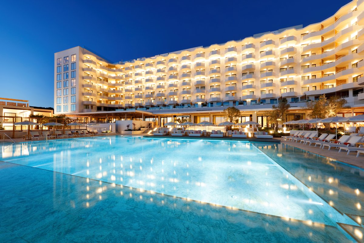 Illuminated pool at twilight with curved resort building, loungers, and palm trees reflected in calm water.