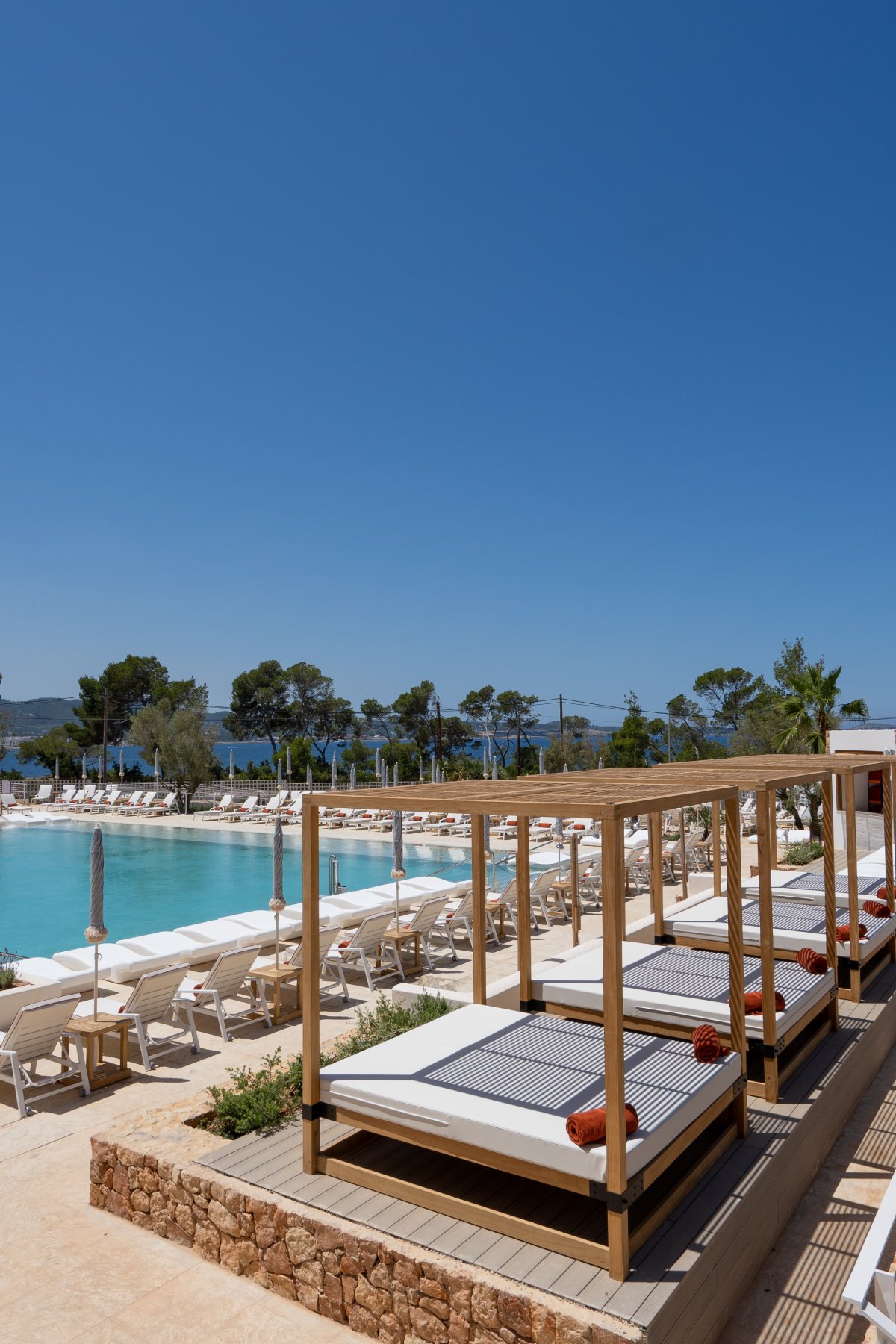 Wooden cabana beds with striped cushions line the pool deck at TRS Ibiza Hotel, overlooking a turquoise pool and distant coastline.