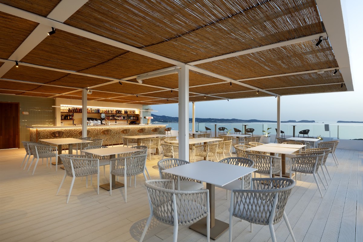 Covered dining terrace with woven wood ceiling, white chairs, and tables overlooking a calm bay with distant islands at TRS Ibiza Hotel.