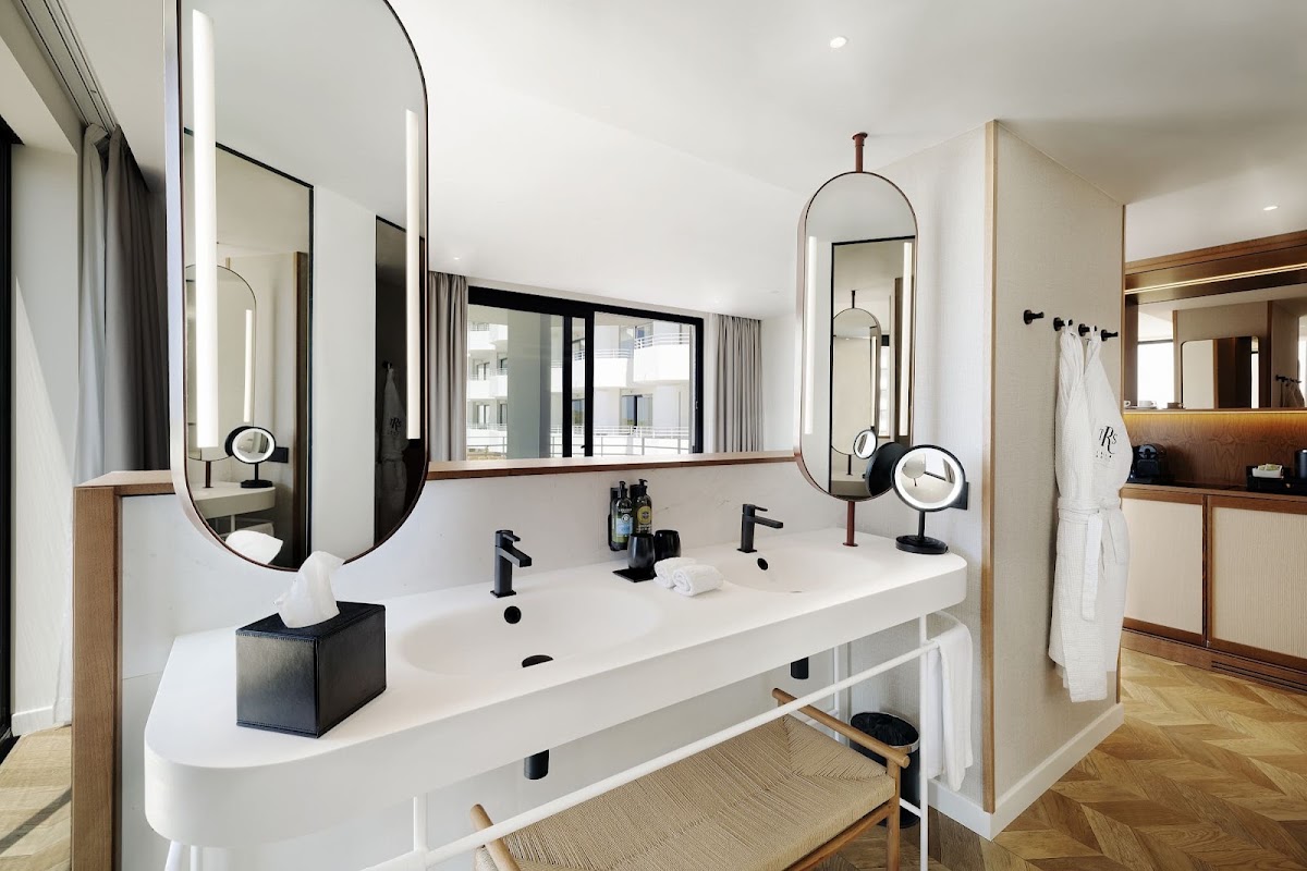 Modern resort bathroom with dual vanity, arched mirrors with lighting, black faucets, and city views through large window.