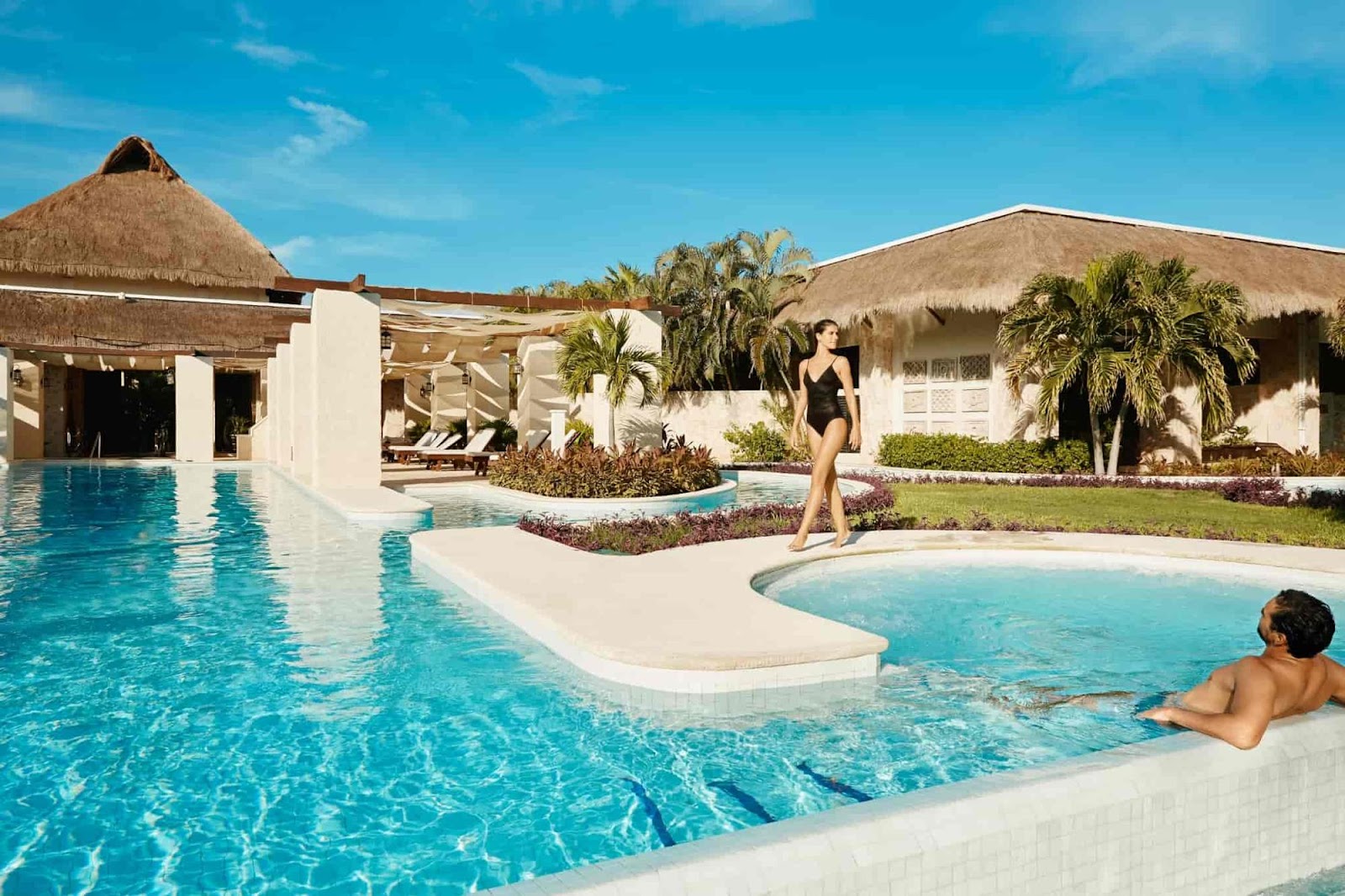 A guest wades in a small pool while another stands nearby at TRS Yucatan Hotel, with thatched-roof structures and white buildings surrounding a larger main pool under clear blue sky.