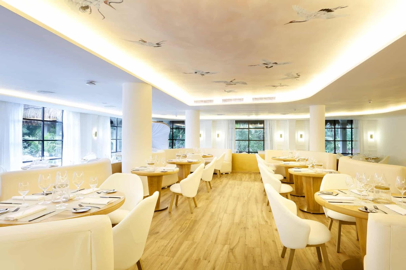 Bright dining room with cream-colored walls, wooden tables, white chairs, and recessed ceiling lights overlooking gardens through large windows.