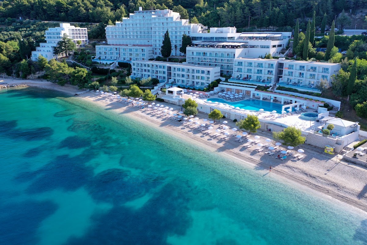 White multi-story resort buildings overlook a turquoise Adriatic beach with rows of loungers, a pool deck, and forested cliffs beyond.