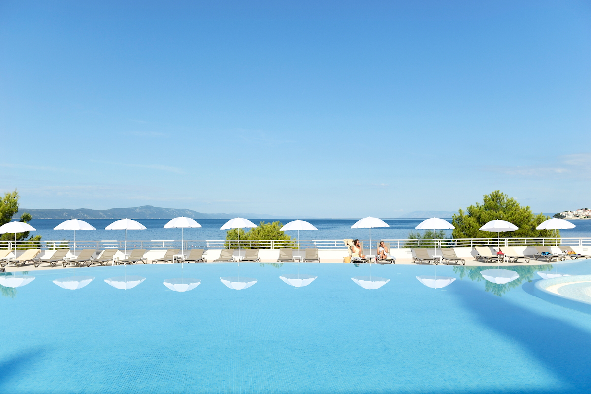 Bright blue pool with white umbrellas and lounge chairs overlooking the Adriatic Sea and distant islands under clear sky.