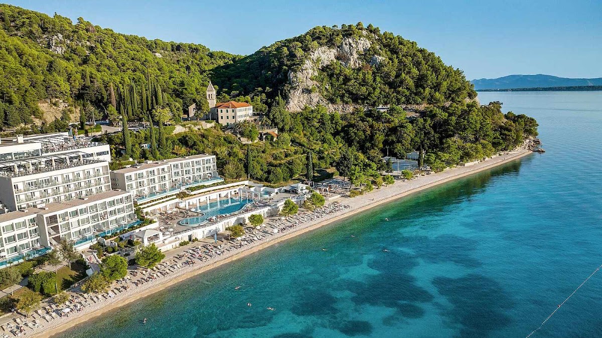 Modern white resort building with pool overlooking a pebble beach and turquoise Adriatic water, surrounded by forested cliffs and a distant church.