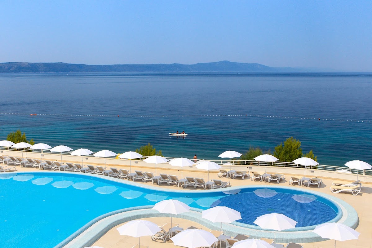 Resort pool with white umbrellas and loungers overlooking a calm turquoise sea, mountains visible across the water.