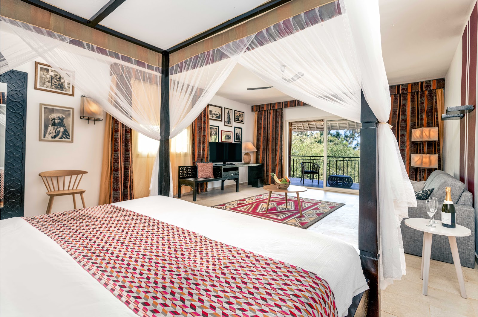 Four-poster bed with white drapes in a resort suite with geometric patterned bedding, sitting area, desk, and balcony overlooking landscaped grounds.