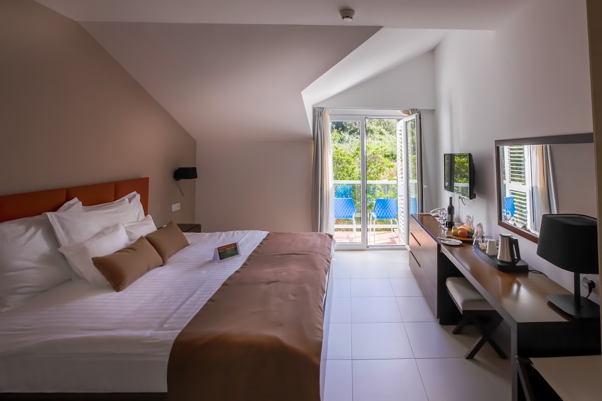 Resort bedroom with queen bed, work desk, and glass doors opening onto a balcony overlooking blue pool and green vegetation.
