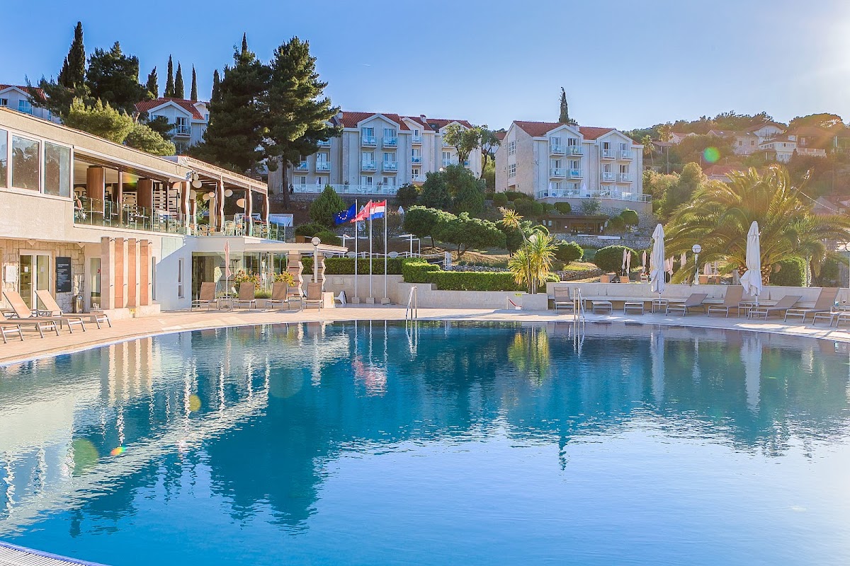 Large swimming pool with white umbrellas and loungers, flanked by a low-rise main building and multi-story residential blocks on a hillside.