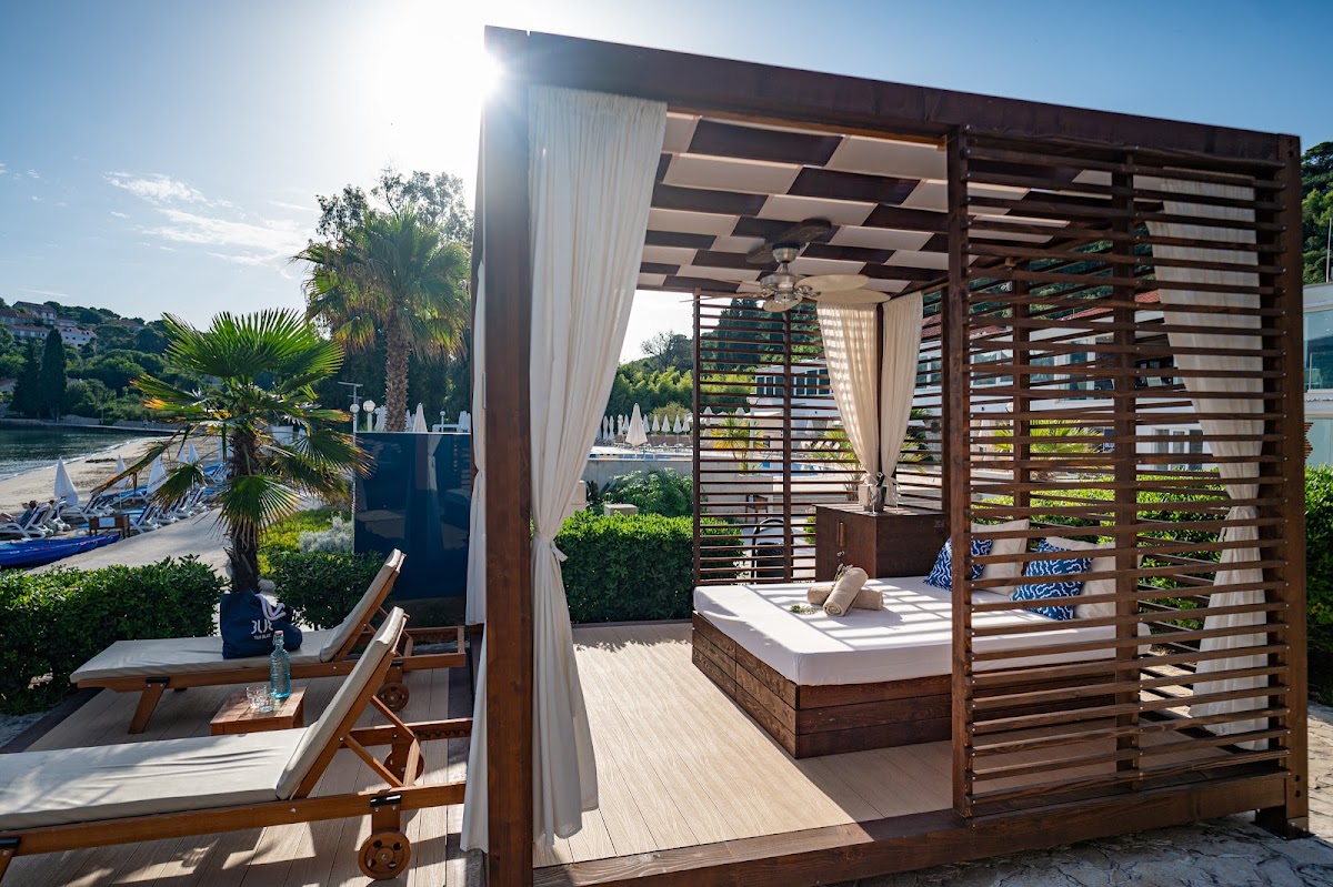Waterfront cabana with slatted wooden frame and white curtains, lounge chairs visible on beach beyond, sailboats moored in water.