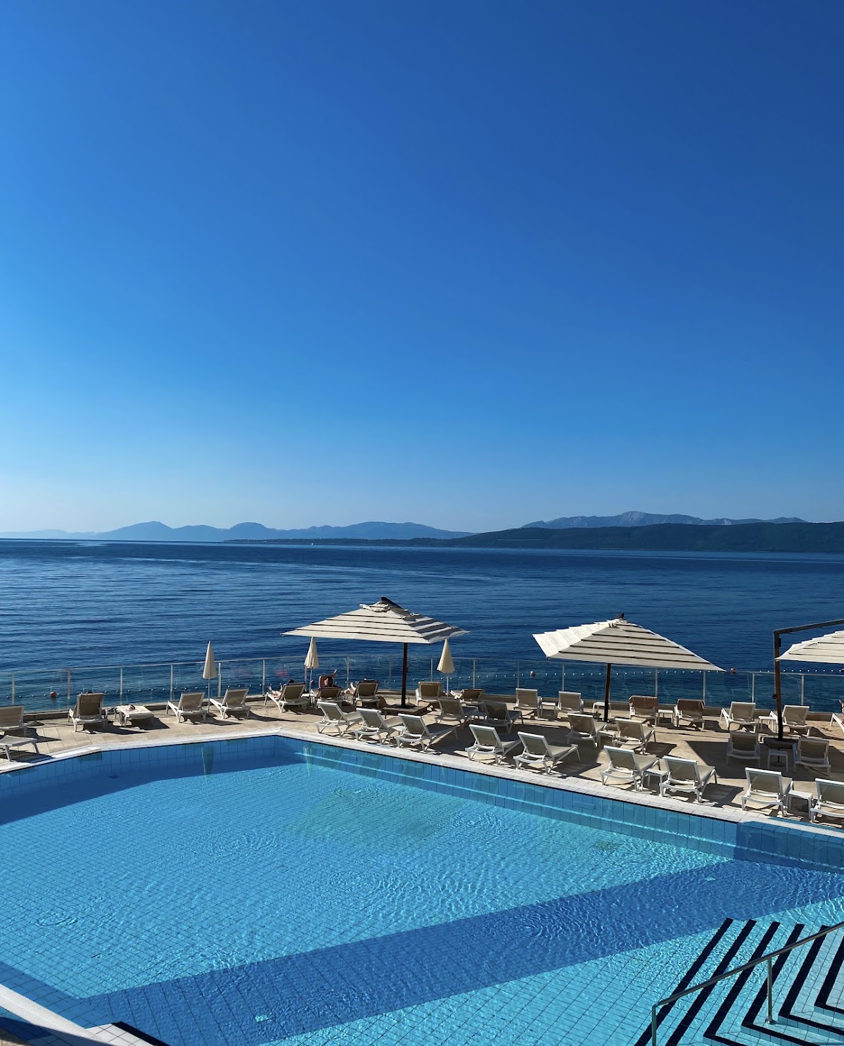 Swimming pool with loungers and striped umbrellas overlooking the Adriatic Sea and distant mountains at TUI BLUE Makarska.
