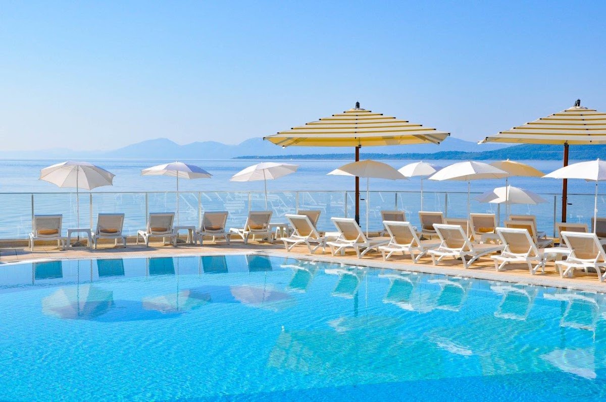 Resort pool with lounge chairs and striped umbrellas overlooking the Adriatic Sea and distant mountains.