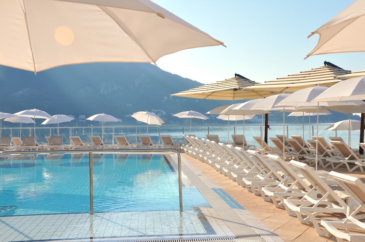Resort pool deck with white lounge chairs and umbrellas overlooking a lake and forested mountains under clear sky.