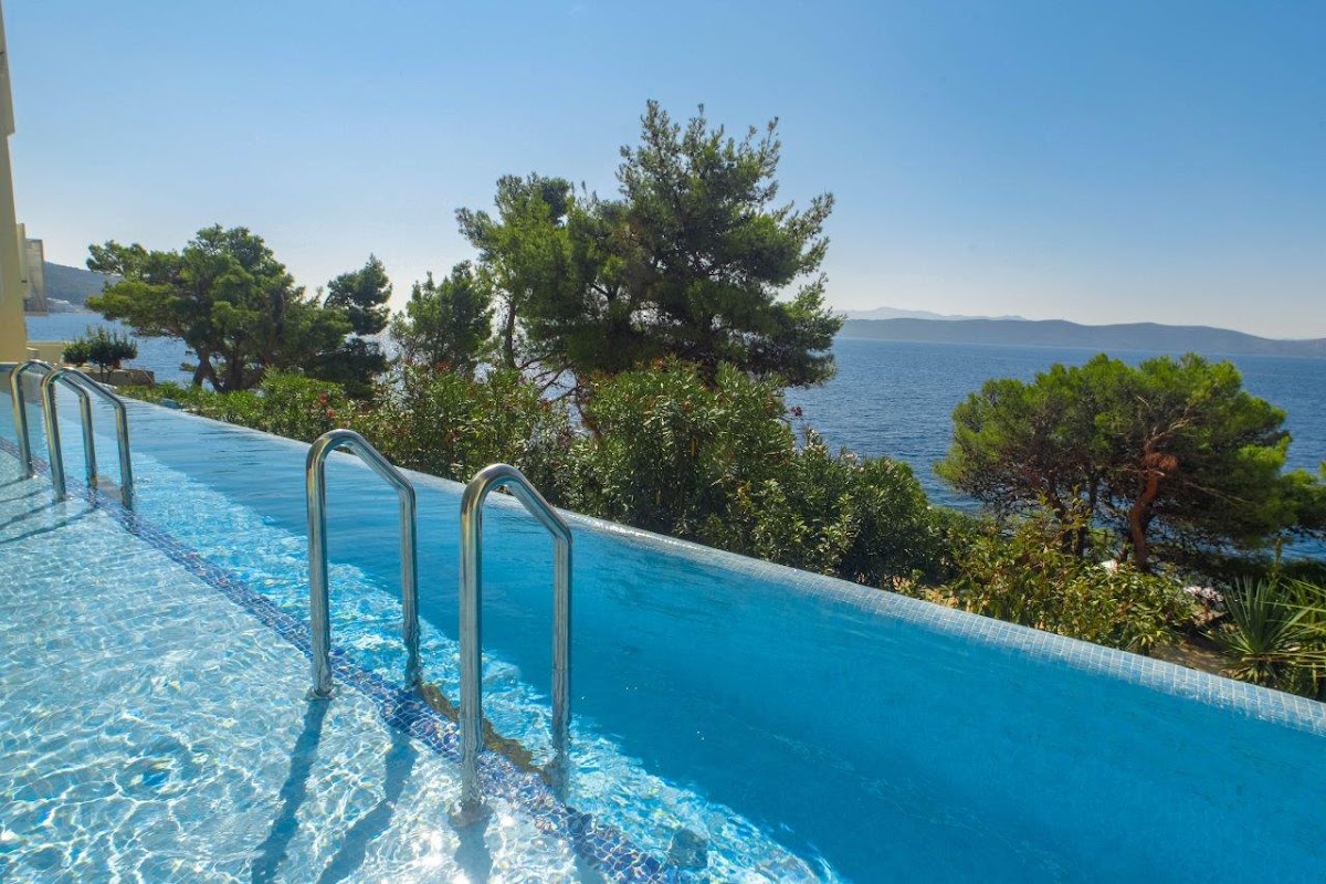 Resort pool with metal railings overlooking the Adriatic Sea and pine trees under clear blue sky.