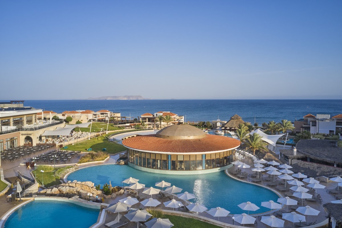 Resort pool complex with white umbrellas, circular roofed building, and Mediterranean Sea beyond, with island visible on horizon.